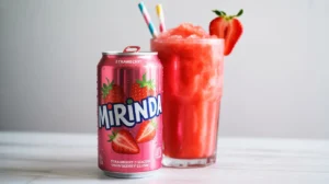 A chilled glass of strawberry slushy made with Strawberry Mirinda beside a pink Mirinda can on a white table.