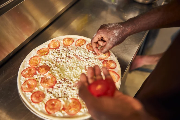 Close-up of a chef’s hands adding sliced tomatoes and cheese on pizza dough in a kitchen.
