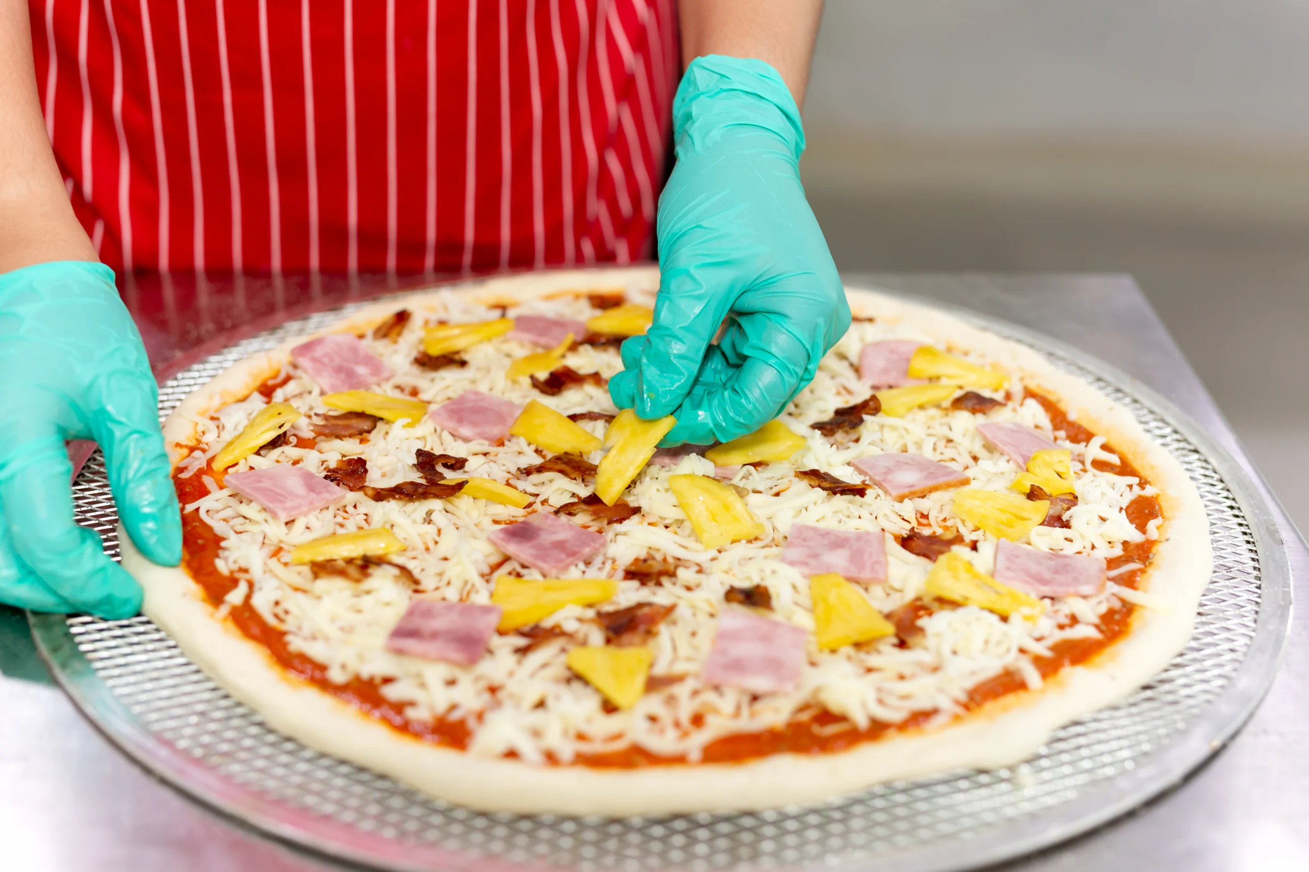 A chef adds pineapple and ham to a Hawaiian pizza one of the most popular pizza topping ideas that blends sweet and savory flavors perfectly.