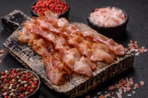 Crispy cooked bacon strips on a rustic wooden board surrounded by bowls of pink salt and mixed peppercorns.