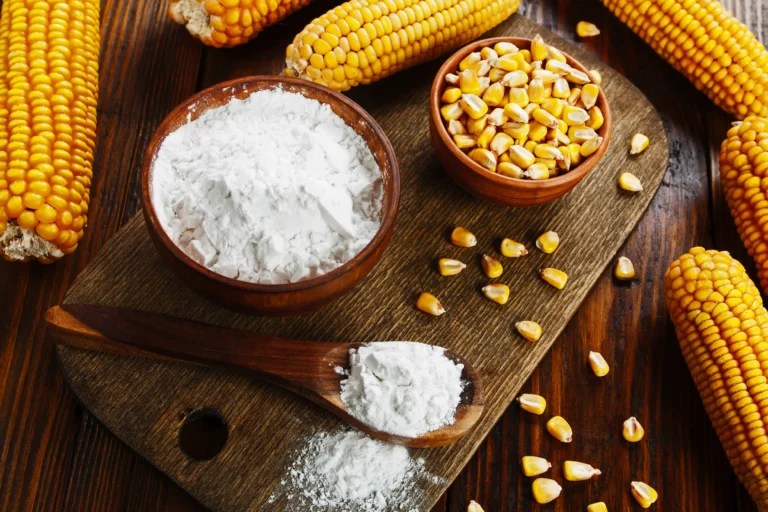 Bowl of white cornstarch and a small bowl of yellow corn kernels on a wooden board, surrounded by whole corn cobs and loose kernels.